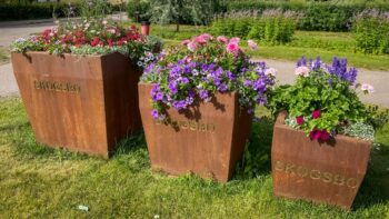 Blomsterplanteringar i stora rostiga krukor med texten Skogsbo på.